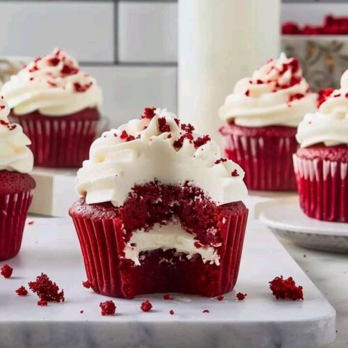 Moist red velvet cupcake with cream cheese frosting and a bite taken out, on marble slab with crumbs scattered.